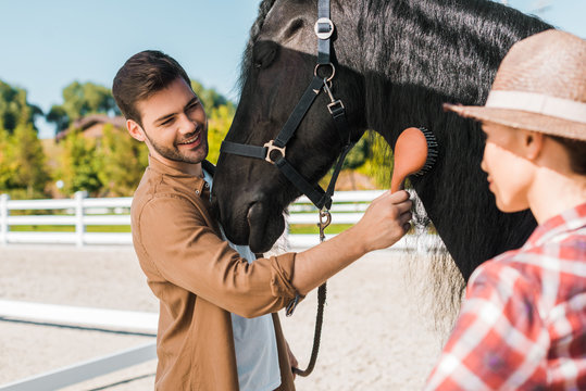 handsome smiling equestrian combing black horse mane at horse club