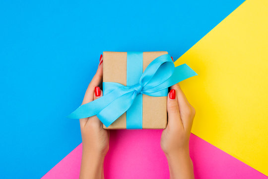 woman hands holding gift wrapped and decorated with blue bow on blue, pink and yellow background with copy space