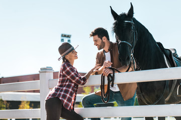 smiling female and male equestrians standing near fence with horse and looking at each other at ranch - obrazy, fototapety, plakaty