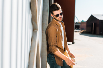 handsome cowboy in sunglasses holding cigarette and leaning on wall at ranch