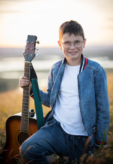 Young teen boy holding acoustic guitar at summer field on sunset and smiling close up