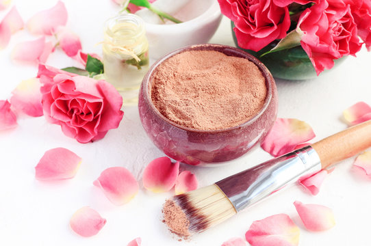 Ceramic bowl of red cosmetic clay for beauty treatment, pink rose petals, aroma oil. Skin care mineral powder mask recipe. 