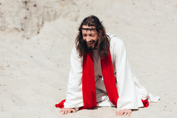 laughing Jesus in robe, red sash and crown of thorns standing on knees and touching sand with hands in desert