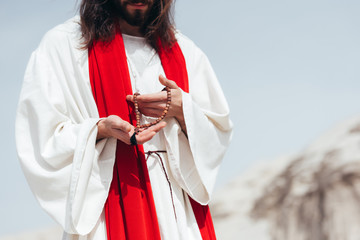 cropped image Jesus with long hair in robe and red sash holding wooden rosary in desert