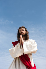 low angle view of Jesus in robe, red sash and crown of thorns holding rosary and praying with...