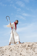 low angle view of Jesus in robe, red sash and crown of thorns walking on sandy hill with staff in...