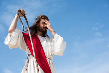 Jesus in robe, red sash and crown of thorns standing with wooden staff in desert and touching...