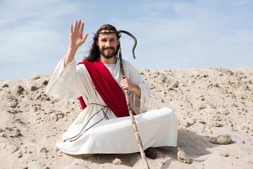 Jesus in robe, red sash and crown of thorns sitting in lotus position on sand in desert and waving hand