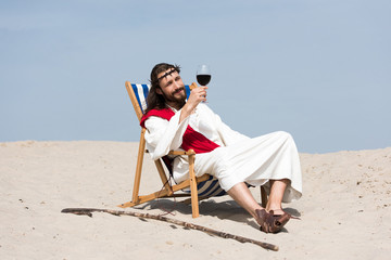 Jesus in robe and red sash resting on sun lounger and looking at glass of red wine in desert