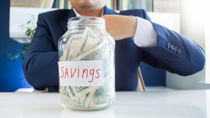 Businessman in blue suit with full glass jar of money savings