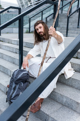 Jesus in robe and crown of thorns sitting on stairs with travel bag and staff, looking at camera