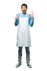 A full-length shot of a Man wearing an apron showing an ok sign with fingers and giving a thumb up gesture with the other hand on isolated background