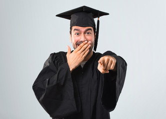 Man on his graduation day University pointing with finger at someone and laughing a lot while covering mouth on grey background