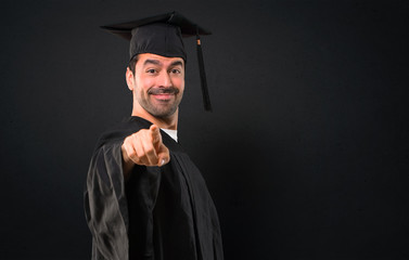 Man on his graduation day University points finger at you with a confident expression on black background