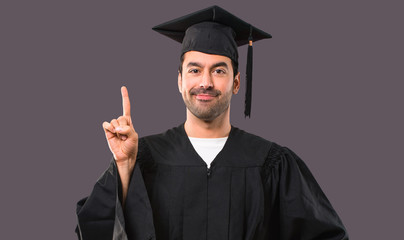 Man on his graduation day University showing and lifting a finger in sign of the best on violet background