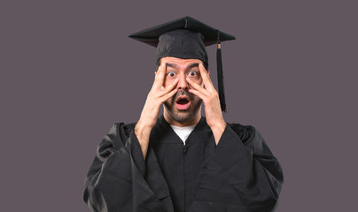 Man on his graduation day University surprised and covering face with hands while looking through fingers on violet background