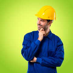Young workman with helmet standing and looking to the side with the hand on the chin on green background