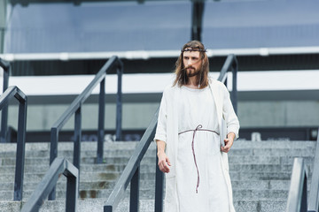 Jesus in robe and crown of thorns walking on stairs with laptop and listening music in city