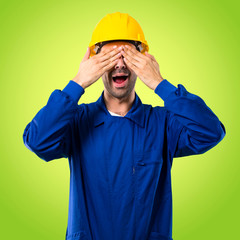 Young workman with helmet covering eyes by hands. Surprised to see what is ahead on green background