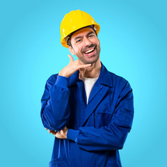 Young workman with helmet making phone gesture and speaking with someone. Call me back sign on blue background