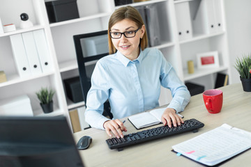 A young girl in glasses sits at a table in the office, holds a pencil in her hand and types the text on the keyboard.