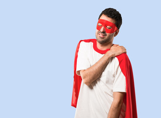 Superhero man with mask and red cape suffering from pain in shoulder for having made an effort on isolated blue background © luismolinero