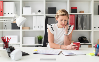 A young blonde girl sits at a computer desk in the office, holds a pencil and a red cup in her hand.
