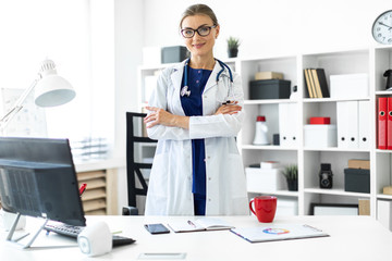 A young girl in a white coat is standing near a table in her office. A stethoscope hangs around her neck.