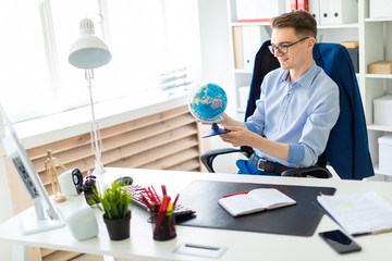 A young man sits in the office at a computer desk and holds a globe in his hands. - obrazy, fototapety, plakaty