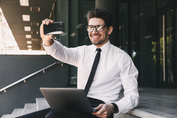 Portrait of cheerful businessman dressed in formal suit sitting outside glass building with laptop, and taking selfie photo on smartphone