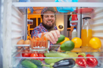 Man standing in front of opened fridge and taking avocado late at night.