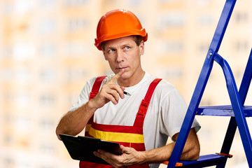 Caucasian foreman checking the object. Worker in a hard hat holding black clipboard and thinking.