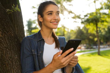 Beautiful woman student walking in park looking aside using mobile phone.