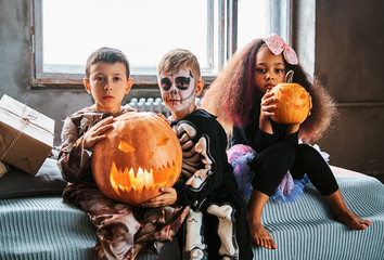 Group of multiracial kids during Halloween party holds pumpkins while sitting on bed in an old...