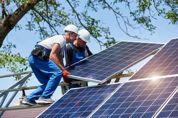 Two young technicians installing heavy solar photo voltaic panel on tall steel platform on green tree background. Exterior solar panel voltaic system installation, dangerous job concept.