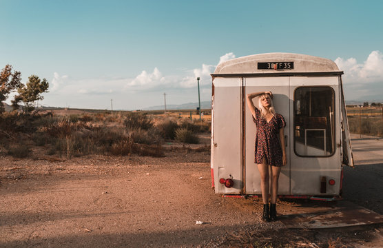 Distant view with copy space of young blonde woman resting on an old van in a sunset of summer blue sky and clouds background
