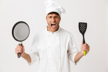 Naklejka premium Angry young chef man standing isolated over white wall background holding frying pan looking camera.