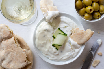 Tzatziki in white ceramic bowl with a cucumber slice and a piece of pita bread next to pita bread, olives and a glass of white wine from above.