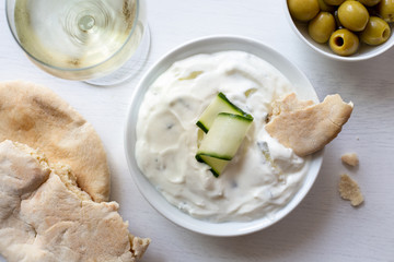 Tzatziki in white ceramic bowl with a cucumber slice and a piece of pita bread next to pita bread, olives and a glass of white wine from above.