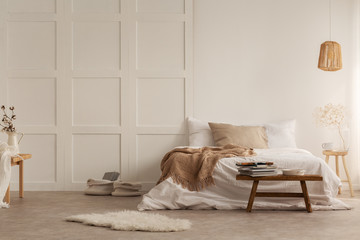 Fur and wooden stool in front of bed with blanket in white bedroom interior with lamp. Real photo