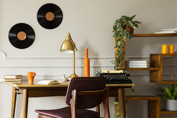 Gold lamp and typewriter on desk in freelancer's interior with vinyl, chair and plants. Real photo
