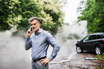 Mature man making a phone call after a car accident, smoke in the background.