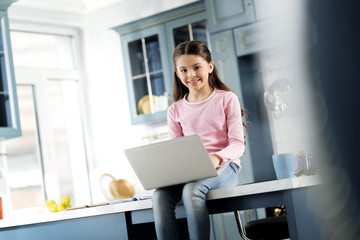 After study. Low angle of satisfied exuberant girl having seat in kitchen and typing on laptop