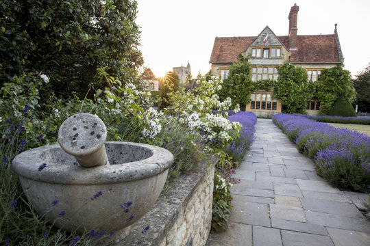 View Of Historic Manor House From Across A Walled Garden With Lawn, Path And Flowerbeds.