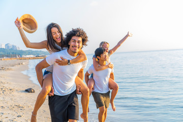 Group of friends walking along the beach, with men giving piggyback ride to girlfriends. Happy young friends enjoying a day at beach