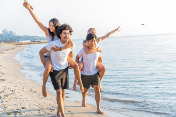 Group of friends walking along the beach, with men giving piggyback ride to girlfriends. Happy young friends enjoying a day at beach