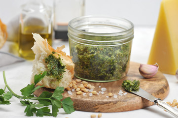 Green pesto sauce in a jar, large salt, garlic clove, pine nuts on a wooden plate. In the frame, a...