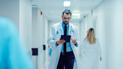 Determined Handsome Doctor Uses Digital Tablet Computer while Walking Through Hospital Hallway....