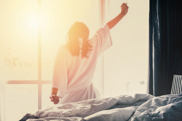 Woman stretching hands on bed after waking up while sitting on bed, entering a day happy and...