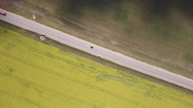 A view from the air dropping down over the highway along which vehicles drive and one car stands amid the yellow and gray fields planted for agriculture by a sunny autumn day